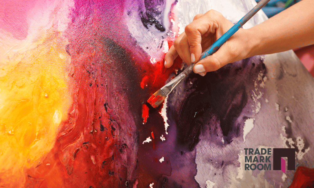 Artist's hand painting vibrant abstract artwork with a brush, featuring a blend of yellow, red, and purple hues, alongside Trademark Room logo.