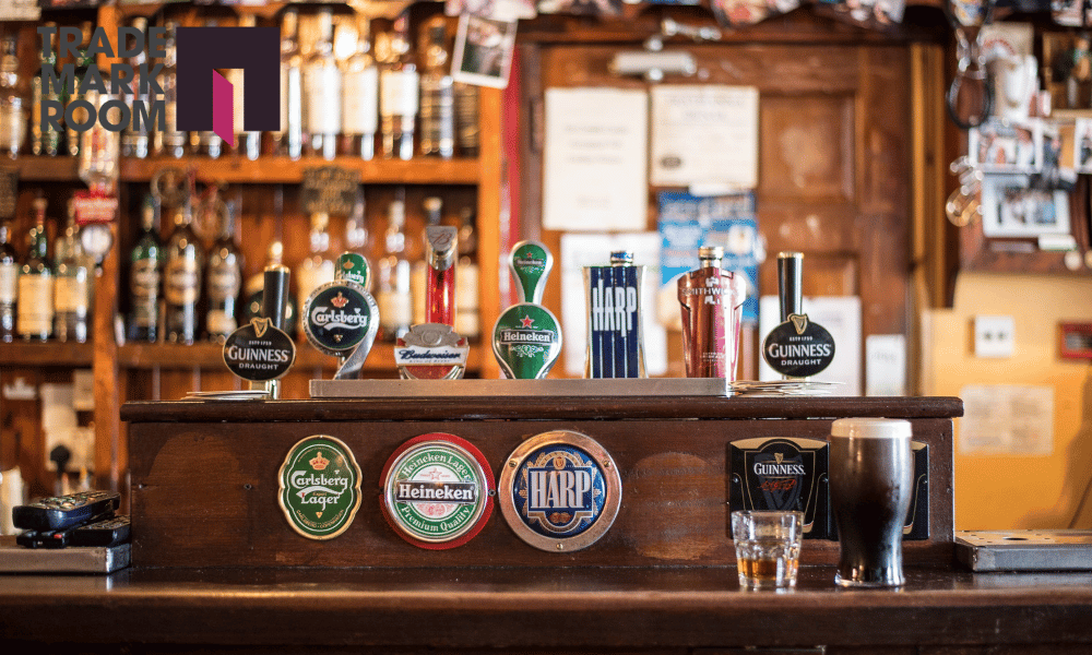 Bar counter featuring various beer taps including Guinness, Heineken, and Carlsberg, with a glass of dark beer and a shot glass, set against a backdrop of liquor bottles, relevant to trademark considerations for bar owners in London.