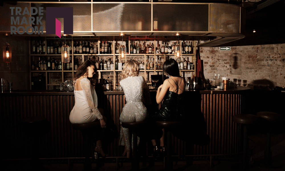 Three women sitting at a bar, showcasing a stylish interior with a well-stocked liquor shelf, highlighting the importance of visual identity for bar owners in London, with TrademarkRoom logo in the corner.