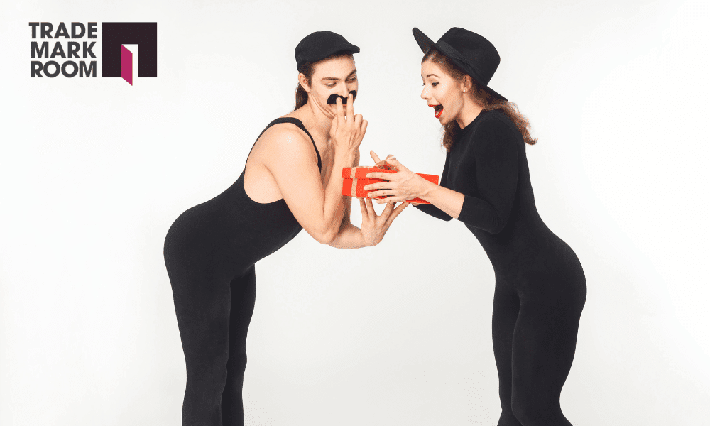 Two individuals in black bodysuits, one with a hat, excitedly exchanging a red gift box, with the Trademark Room logo in the top left corner, illustrating themes of parody and brand engagement in digital culture.