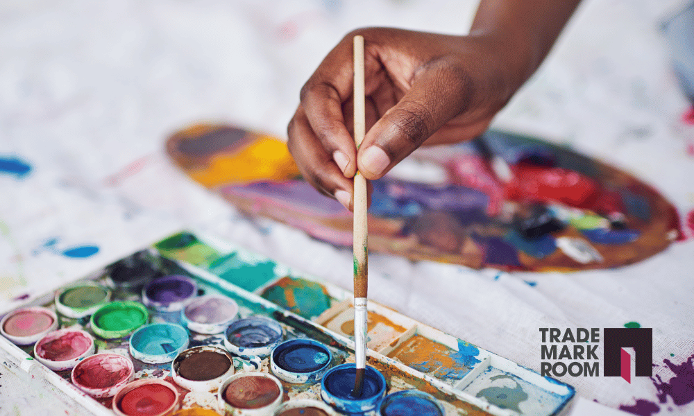 Hand holding paintbrush above a palette of vibrant watercolours, illustrating artistic creation and the importance of trademark protection for artists.