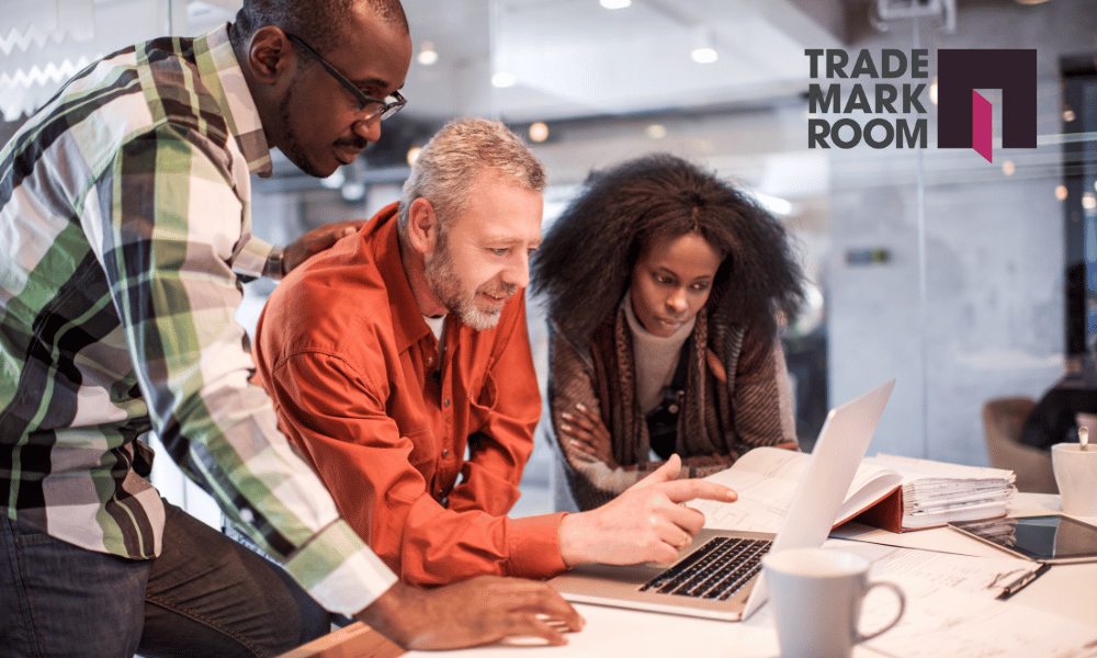 Three professionals collaborating at a table, reviewing documents and a laptop, with Trademarkroom logo visible, emphasising intellectual property strategy and legal guidance for small businesses.