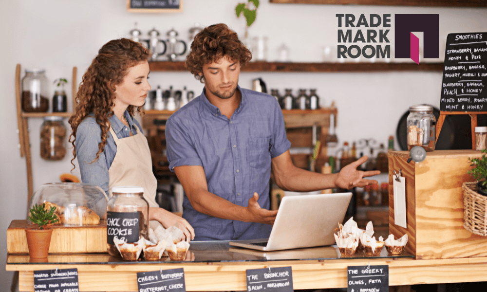Barista and customer discussing business at a café counter with trademark registration materials, featuring "Trademark Room" logo and various baked goods.